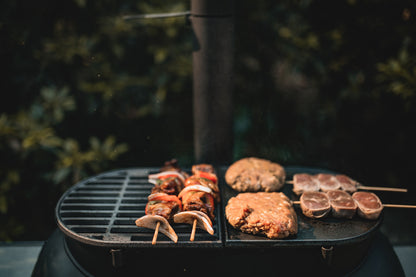 Skewers of vegetables and meat, along with two burger patties and three round meat skewers, cook outdoors on the Ozpig Big Pig Flat Iron BBQ Plate by Ozpig, set against a leafy backdrop that enhances the wood-fired BBQ experience.