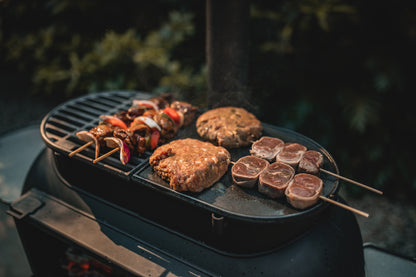 A close-up of assorted meats and vegetable skewers sizzling on the Ozpig Big Pig Flat Iron BBQ Plate by Ozpig, with blurred greenery in the background.