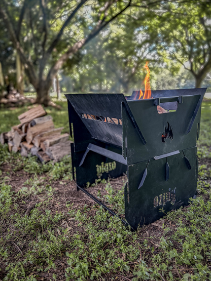 An Ozpig 3-in-1 Flat Pack Fire Pit, with flames burning inside its black metal frame, stands on the grass next to a stack of firewood, surrounded by trees and greenery in the background.