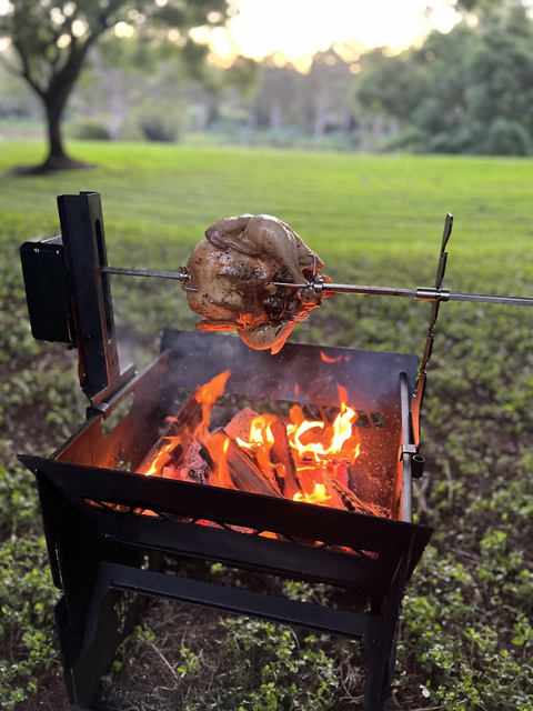 A whole chicken is roasting outdoors on the Ozpig Fire Pit Rotisserie Kit by Ozpig, turning gently above an open flame grill and soaking up a rich wood-fired flavour, with green grass and trees in the background at sunset.