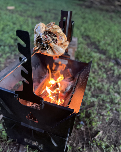 The Ozpig Fire Pit Rotisserie Kit by Ozpig lets you roast a whole chicken on a rotisserie spit over an open flame in a sturdy metal grill, delivering mouthwatering wood-fired flavour—ideal for outdoor cooking with green grass and blurred trees as your scenic backdrop.