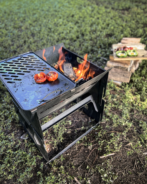 An Ozpig Fire Pit Solid/Grill Cooking Plate by Ozpig is used to cook sliced tomatoes outdoors over an open fire, providing even heat distribution thanks to its heavy-duty steel construction. In the background, a stack of wooden planks and a plate of food are placed on the grass.