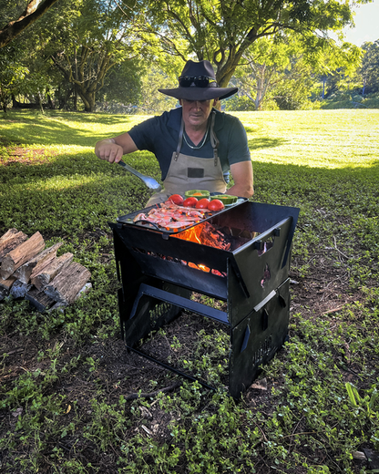 A person wearing a wide-brimmed hat and apron grills meat and vegetables on an Ozpig Fire Pit using the Ozpig Fire Pit Solid/Grill Cooking Plate by Ozpig, in a grassy, sunlit park with trees in the background.