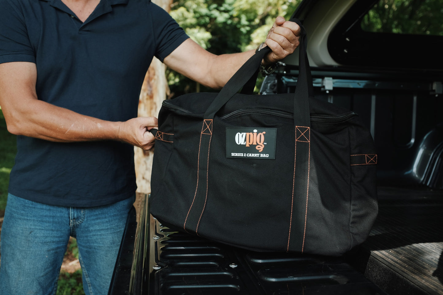 A person places the Ozpig Series 2 Heavy Duty Storage Bag by Ozpig—featuring durable canvas construction, orange stitching, and internal pockets—into the back of a pickup truck outdoors.