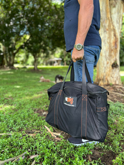 A person stands on grass holding a large black Ozpig Series 2 Heavy Duty Storage Bag by the handles, crafted from heavy duty canvas, with trees and greenery in the background.