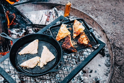 Slices of bread are being cooked over a campfire on a grill. Some are in a pre-seasoned cast iron skillet, while others are browned on the Campfire 24cm Square Grill Frypan with Folding Handle, with ashes and logs visible around the fire pit.