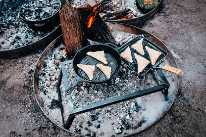 Slices of bread are being toasted in two Campfire 24cm Square Grill Frypans with folding handles over an open campfire on a metal grate, surrounded by ash and logs in a fire pit outdoors.