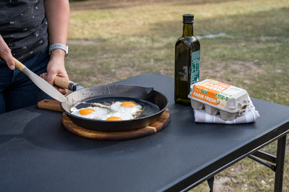 A person cooks three sunny-side-up eggs in the Campfire Round Frying Pan - Solid Handle 30cm on a black table outdoors, with a bottle of olive oil and a carton of farm fresh eggs nearby.