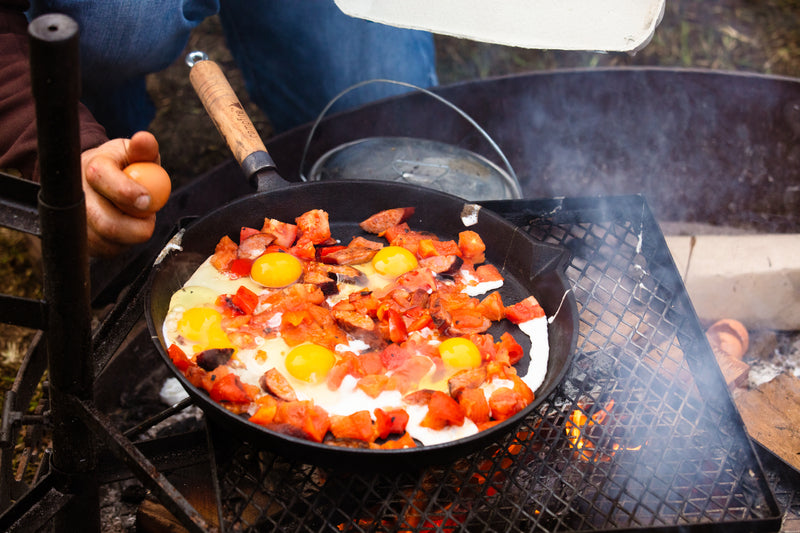 A person cooking eggs and chopped vegetables in the Campfire Round Frying pan - Solid Handle 30cm over an open fire, with one hand holding an egg ready to crack into the 30cm skillet. Smoke rises from the flames below the grate.