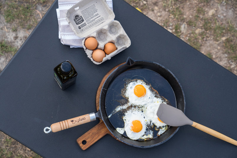 A pre-seasoned Campfire Round Frying Pan - Solid Handle 30cm is shown on a black table with two sunny-side-up eggs inside, accompanied by a spatula, an open egg carton with eggs, a towel, and a dark bottle. This versatile frypan features a solid handle for comfortable use and easy handling.