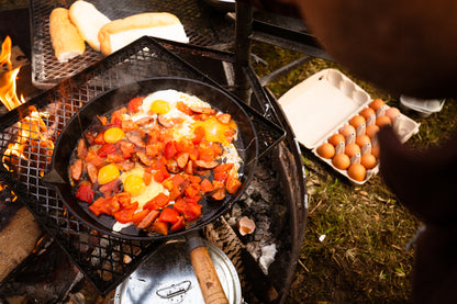 A Campfire 25cm Round Frying Pan with a solid handle sizzles with eggs, sausage, and tomatoes over an outdoor fire; bread toasts beside it while a carton of eggs rests on the grass nearby.