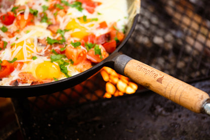 A close-up of the Campfire 25cm Round Frying Pan Solid Handle, pre-seasoned and filled with eggs, tomatoes, cheese, and herbs sizzling over a campfire grill. The skillet includes a durable removable wood handle engraved with “Campfire” and a flame logo.