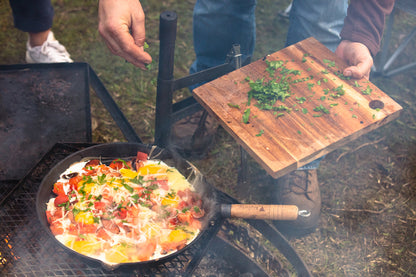 A person sprinkles chopped herbs from a wooden board onto a skillet of eggs, tomatoes, and cheese sizzling in a Campfire 25cm Round Frying Pan Solid Handle over a campfire outdoors.