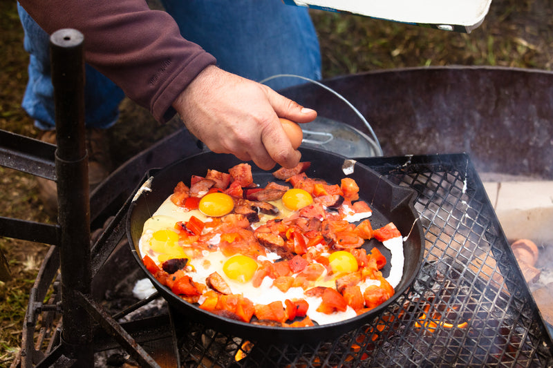 A person cracks an egg into the Campfire 25cm Round Frying Pan with Solid Handle over a campfire. The skillet, filled with eggs, chopped tomatoes, and meat, cooks on a metal grate outdoors.