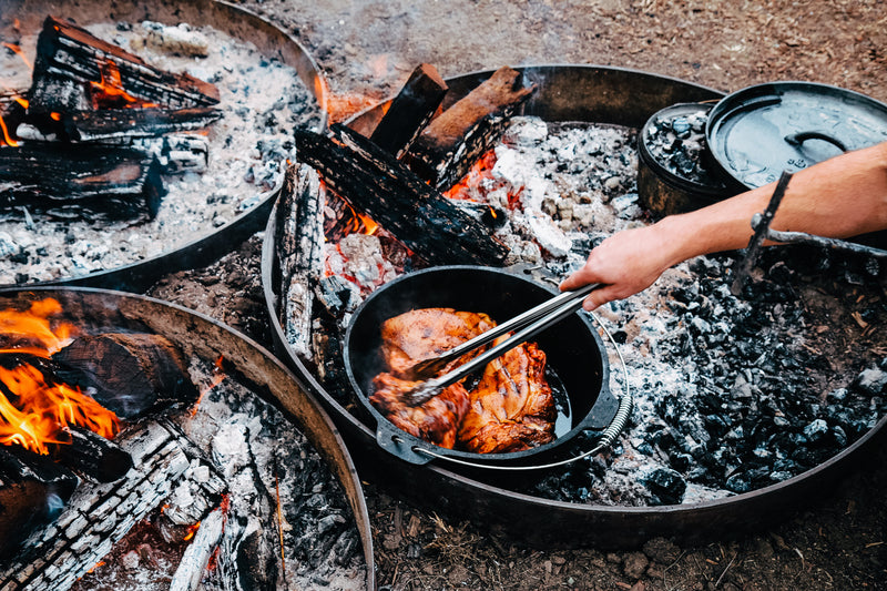 A person uses tongs to cook meat in the Campfire 9 Quart Camp Oven over a campfire, surrounded by burning wood and ash. Other pots and fire rings are visible nearby, capturing the rustic charm of outdoor cooking in a scenic setting.