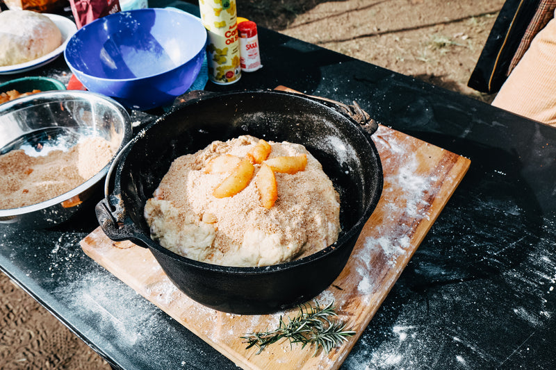 A 9 Quart Camp Oven by Campfire in black cast iron sits on the table, filled with dough topped with breadcrumbs and sliced apples. Mixing bowls, flour, and rosemary are arranged nearby, surrounded by assorted ingredients and cooking tools—ideal for outdoor cooking or making a tasty treat by the campfire.