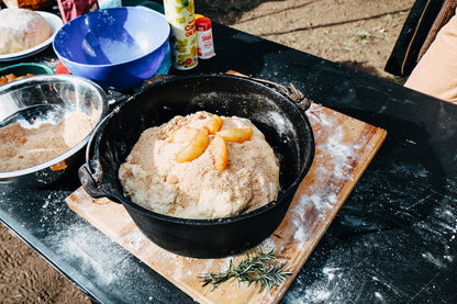 A 9 Quart Camp Oven by Campfire in black cast iron sits on the table, filled with dough topped with breadcrumbs and sliced apples. Mixing bowls, flour, and rosemary are arranged nearby, surrounded by assorted ingredients and cooking tools—ideal for outdoor cooking or making a tasty treat by the campfire.