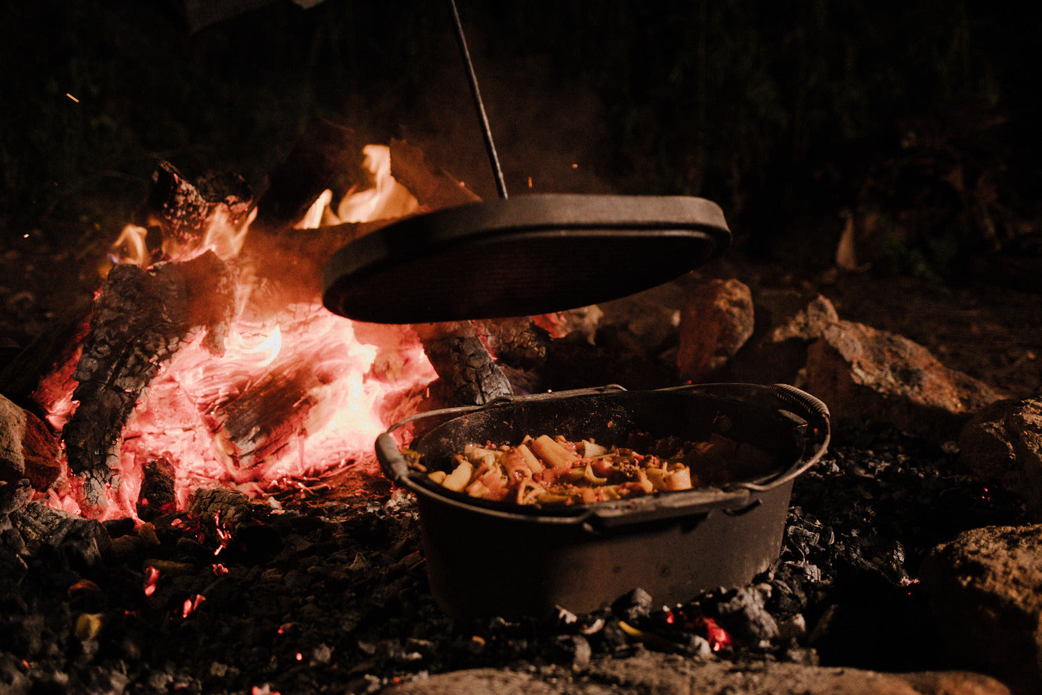 A Campfire 10 Quart Oval Camp Oven filled with pasta and vegetables sits over hot coals by a campfire, its lid being lifted in a nighttime outdoor setting.