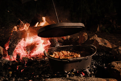 A Campfire 10 Quart Oval Camp Oven filled with pasta and vegetables sits over hot coals by a campfire, its lid being lifted in a nighttime outdoor setting.