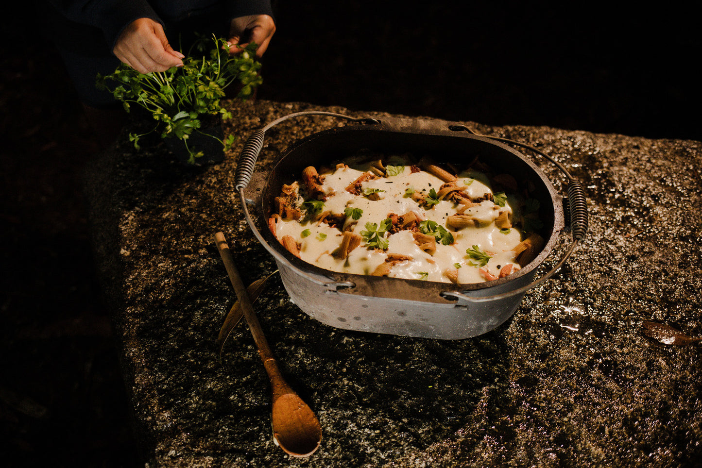 A person holds fresh herbs next to a rustic Campfire 10 Quart Oval Camp Oven filled with baked pasta topped with melted cheese, placed on a stone surface with two wooden spoons nearby.