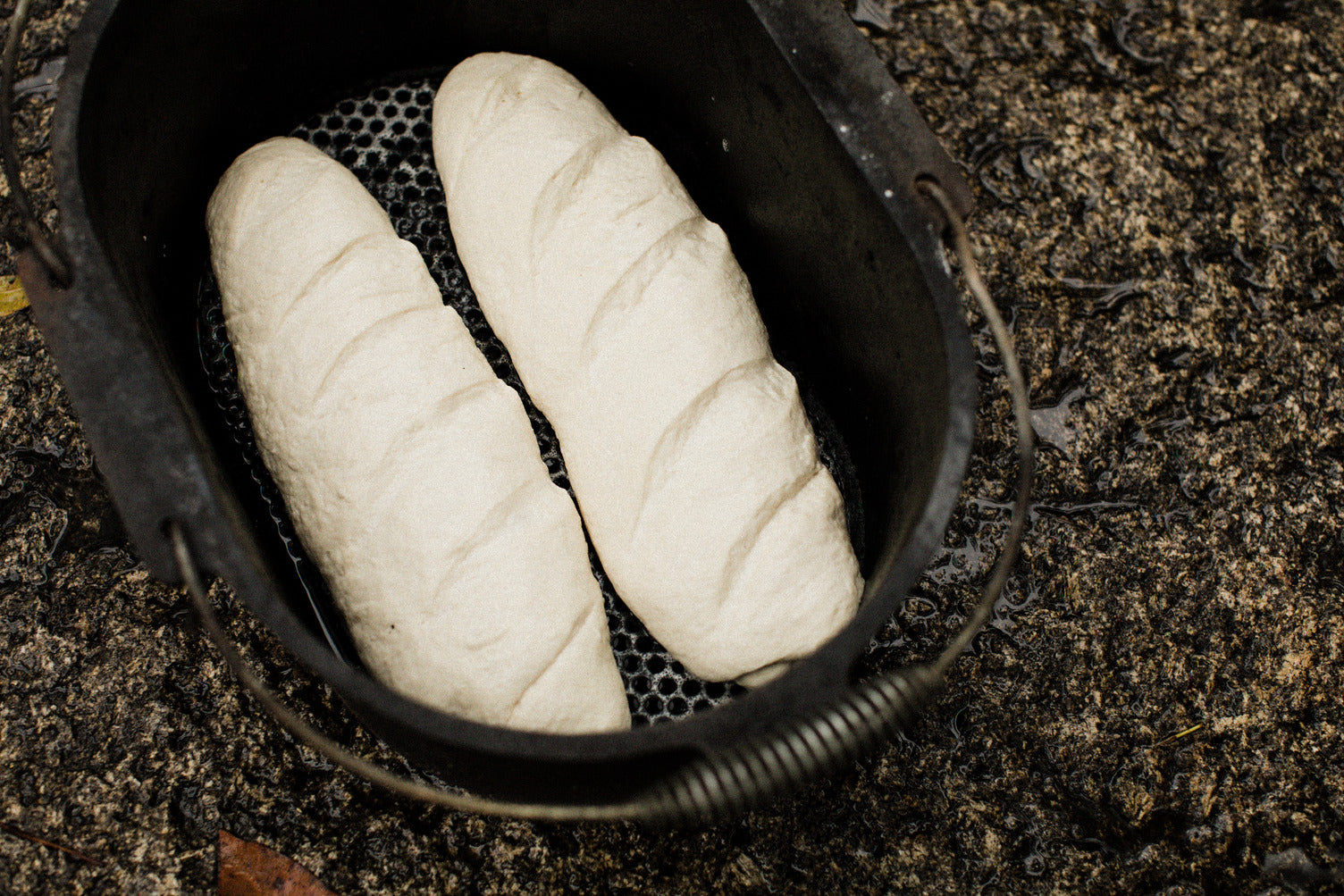 Two unbaked loaves of bread dough rest side by side in a Campfire 10 Quart Oval Camp Oven, its black cast iron construction positioned on a rough, wet outdoor surface—making it perfectly suited for campfire cooking or use as a camp oven.