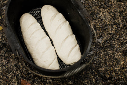 Two unbaked loaves of bread dough rest side by side in a Campfire 10 Quart Oval Camp Oven, its black cast iron construction positioned on a rough, wet outdoor surface—making it perfectly suited for campfire cooking or use as a camp oven.