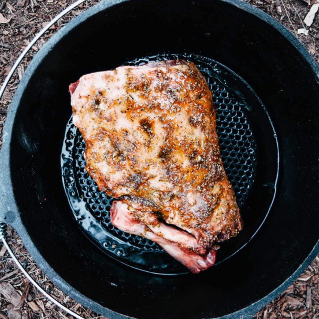 A seasoned, cooked piece of meat sits in a black cast iron pan atop the Campfire Large Trivet - Suits 9 Quart, viewed from above, with specks of herbs and juices visible on the surface—a perfect example of campfire cooking.