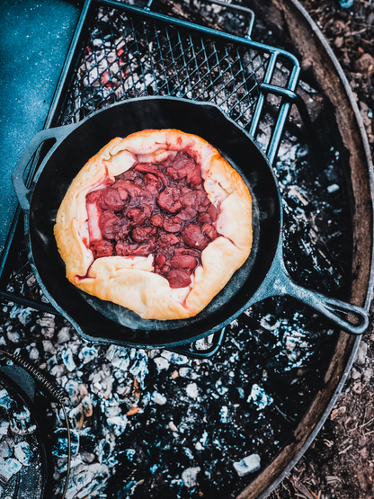 A rustic cherry galette sizzles in the Campfire 30cm Cast Iron Skillet over an outdoor fire pit, surrounded by ashes and a grill grate—perfect for campfire cooking.