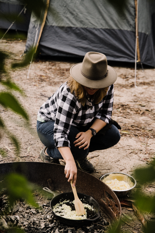 Wearing a plaid shirt and hat, a person cooks food in the 30cm Cast Iron Skillet by Campfire over an open fire, surrounded by nature with tents and a bowl of ingredients nearby.