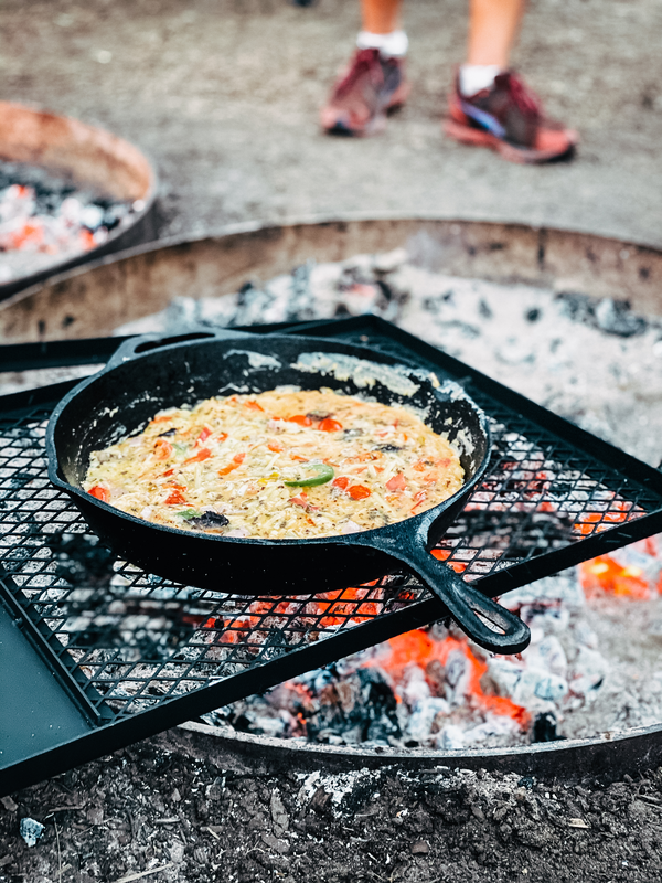 The Campfire 30cm Cast Iron Skillet, filled with a vegetable dish, cooks over an open campfire on a grill grate. Glowing embers illuminate the scene, while a person in sneakers stands in the background, highlighting the art of campfire cooking.