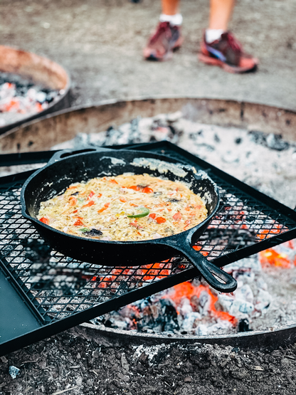 The Campfire 30cm Cast Iron Skillet, filled with a vegetable dish, cooks over an open campfire on a grill grate. Glowing embers illuminate the scene, while a person in sneakers stands in the background, highlighting the art of campfire cooking.