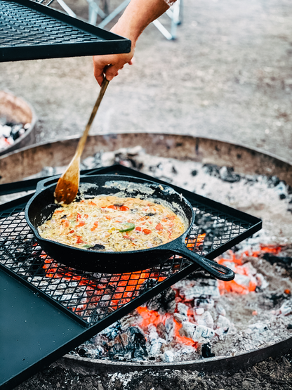 A person enjoys campfire cooking, stirring food in the durable 30cm Cast Iron Skillet by Campfire over open flames and glowing embers. The skillet rests securely on a metal grate outdoors, capturing the rustic spirit of outdoor meals.