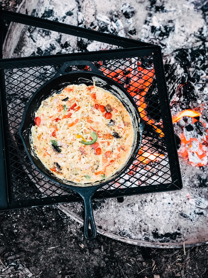 The Campfire 30cm Cast Iron Skillet, filled with a vibrant vegetable omelet, rests on a grill above a campfire, surrounded by ash and glowing embers—perfectly capturing the essence of rustic campfire cooking.