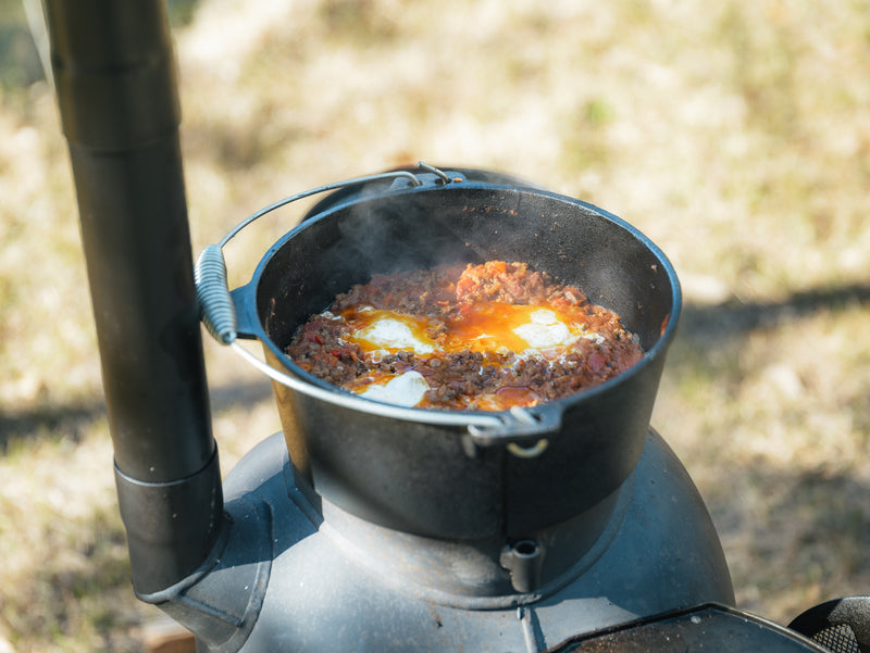 A Campfire 4.5 Quart Camp Oven sits atop an outdoor stove, filled with a simmering tomato-based stew featuring visible eggs. Steam rises from the hot dish, while softly blurred grass in the background captures the essence of outdoor cooking.
