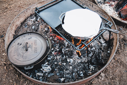 Surrounded by a fire ring outdoors, hot coals heat the Campfire 4.5 Quart Camp Oven and a grill grate with a skillet covered in foil, creating the perfect setting for outdoor cooking.