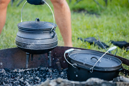 A person lifts a black Campfire 8L Cast Iron Potjie Pot over a campfire pit with charcoal, next to another lidded cast iron pot, showcasing classic outdoor cooking with grass in the background.