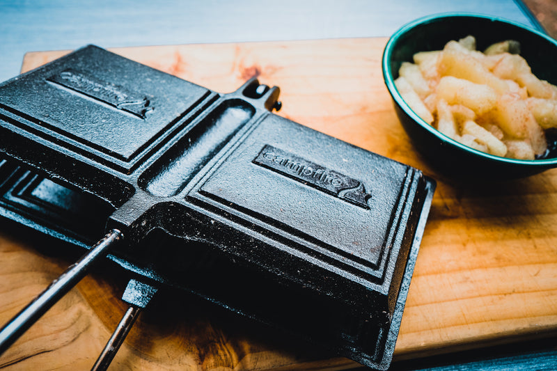 A Campfire Jaffle Iron Jumbo Double, crafted from black cast iron and perfect for preparing campfire meals, is placed on a wooden cutting board beside a small bowl of cooked, sliced apples.