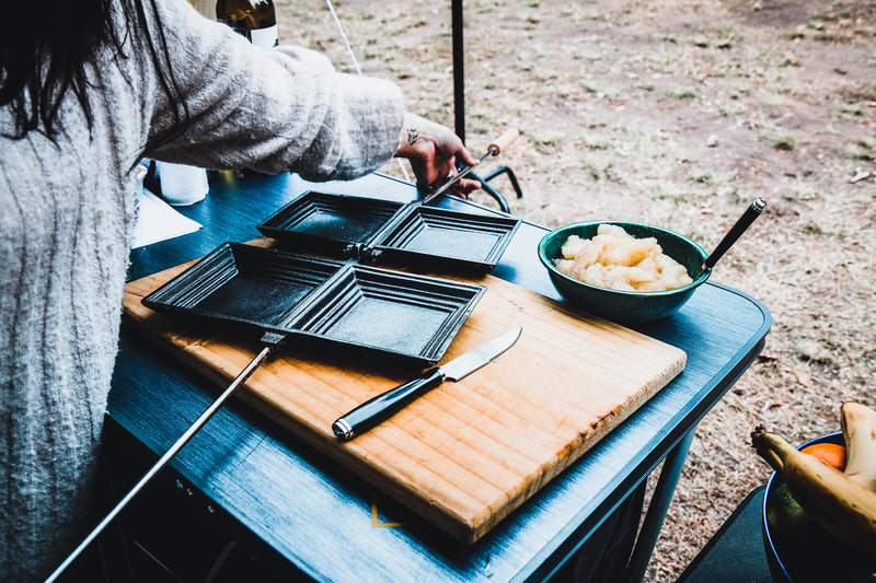 A person prepares food outdoors, with a Campfire Jaffle Iron Jumbo Double open next to a knife on a wooden cutting board, and a bowl of cut fruit on a blue table.