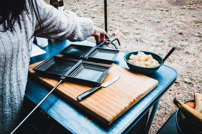 A person prepares food outdoors, with a Campfire Jaffle Iron Jumbo Double open next to a knife on a wooden cutting board, and a bowl of cut fruit on a blue table.