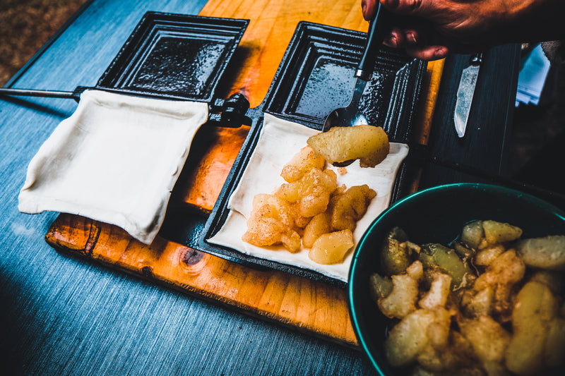 A hand spoons apple filling onto square dough pieces arranged on black Campfire Jaffle Iron Jumbo Double pie irons, with a bowl of extra filling nearby on the wooden surface—a delicious campfire meal in the making.