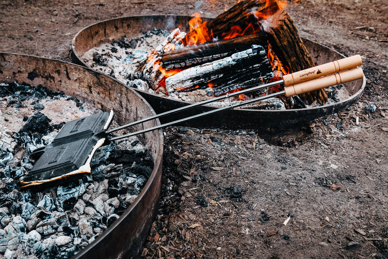 Two round fire pits: one with burning wood, the other with a Campfire Jaffle Iron Jumbo Double sandwich press containing toasted sandwiches, resting on hot coals. The ground is dirt covered with ashes and bits of wood—an inviting scene for classic campfire meals.