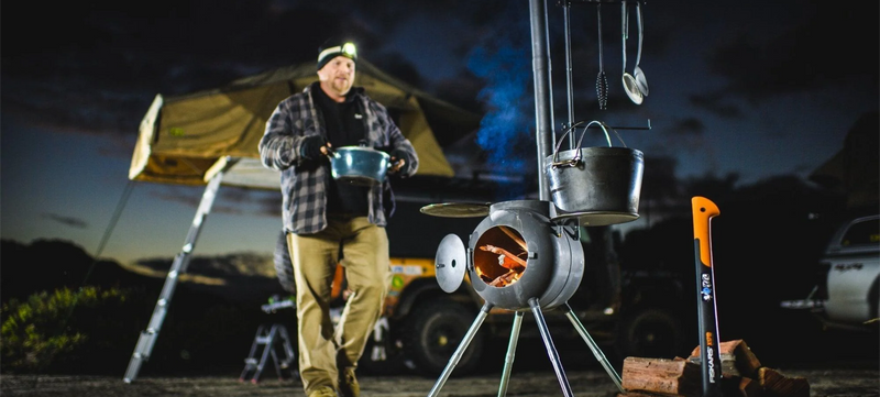 A man wearing a headlamp carries a pot near a camp stove with burning wood, a kettle, and pans hanging above. There’s an axe, firewood, and a rooftop tent with a ladder in the background, all at dusk.