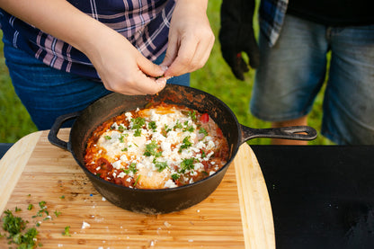 Outdoors, someone sprinkles fresh herbs onto a cheesy vegetable dish in a Campfire Cast Iron Combo Cooker 3.2 Quart, which rests on a wooden cutting board as another person stands nearby.