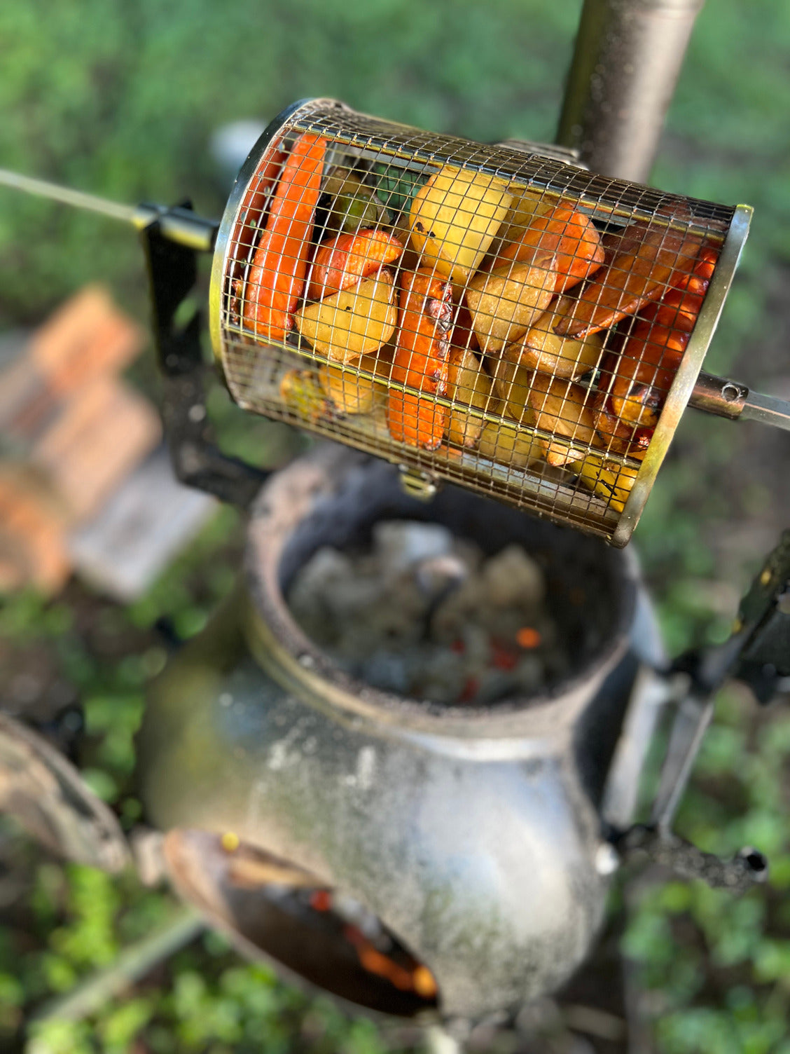 An Ozpig Rotisserie Basket Small from Ozpig, filled with potatoes and carrots, hangs over a black outdoor fire pit with hot coals glowing beneath, set in a grassy outdoor area.