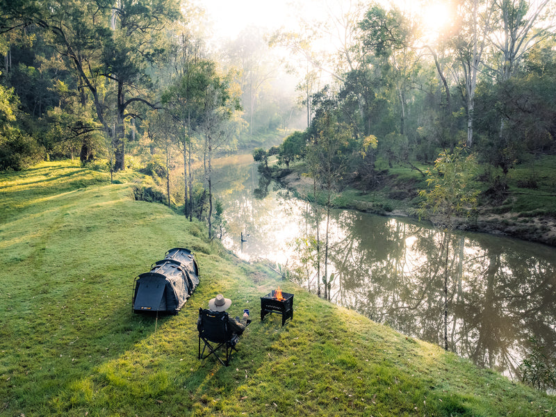 A person relaxes in a camping chair beside the Ozpig 3-in-1 Flat Pack Fire Pit near a tent, enjoying views of a calm river bordered by trees and lush greenery on a sunny morning.