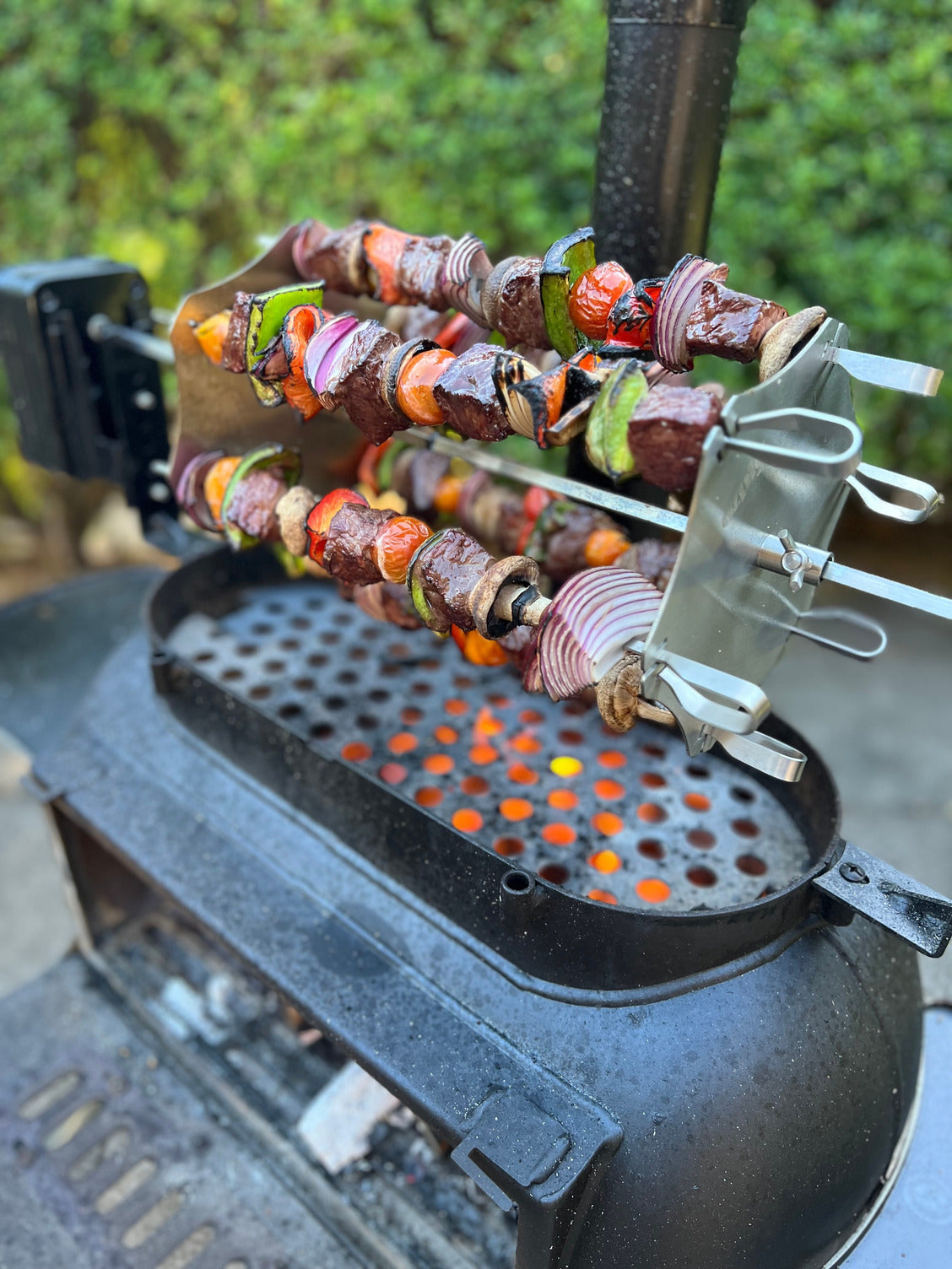 Colorful meat, bell pepper, onion, and zucchini skewers grill over hot charcoal on an Ozpig Kebab Rotisserie Set by Ozpig, using stainless steel skewers with a blurred green background.