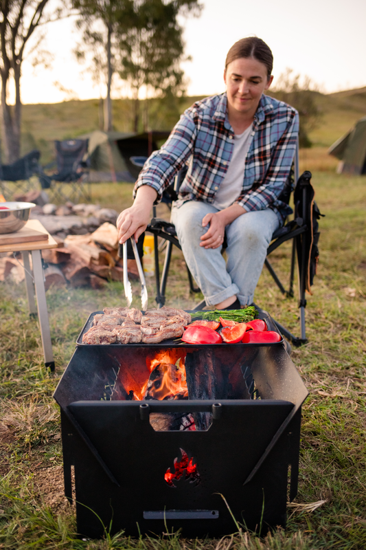 Wearing a plaid shirt and jeans, a person grills meat and vegetables over the Ozpig 3-in-1 Flat Pack Fire Pit by a campfire, with tents and trees in the background, creating a cozy campsite atmosphere.