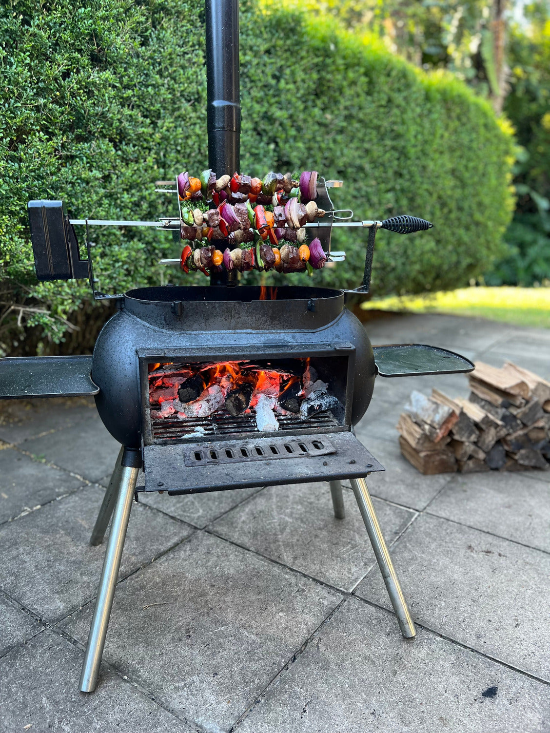 The Ozpig Kebab Rotisserie Set by Ozpig holds stainless steel skewers of colorful vegetables above burning wood in a black outdoor grill, surrounded by stacked logs on a tiled patio with green hedges behind.