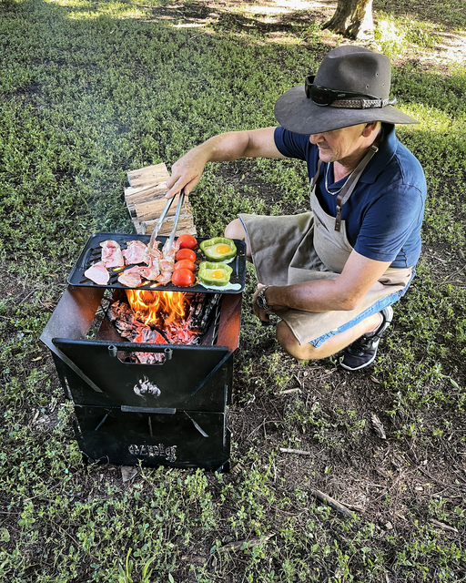 A man kneels on the grass, using tongs to grill meat and vegetables over an open flame on the Ozpig 3-in-1 Flat Pack Fire Pit, with wood stacked nearby.