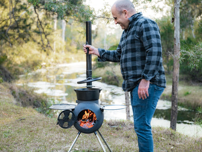 A man in a plaid shirt stands outdoors by a small wood-burning stove with a visible fire, lifting the lid off his Campfire 4.5 Quart Camp Oven. Trees, grass, and a creek are in the background, evoking authentic outdoor cooking.